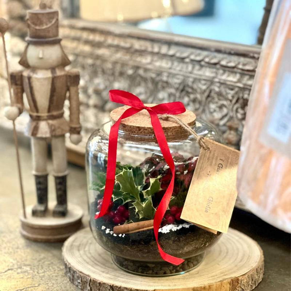 Terrarium natalizio in barattolo di vetro con agrifoglio, bacche rosse e nastro rosso decorativo.