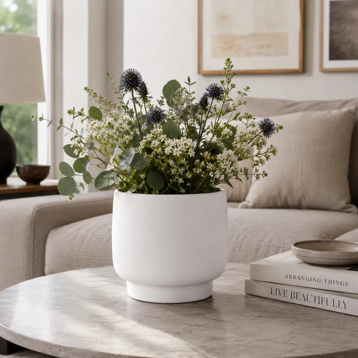 Vaso Sweet bianco con composizione floreale composta da eucalipto, fiorellini bianchi e cardi blu, posizionato su tavolino in un elegante soggiorno contemporaneo.