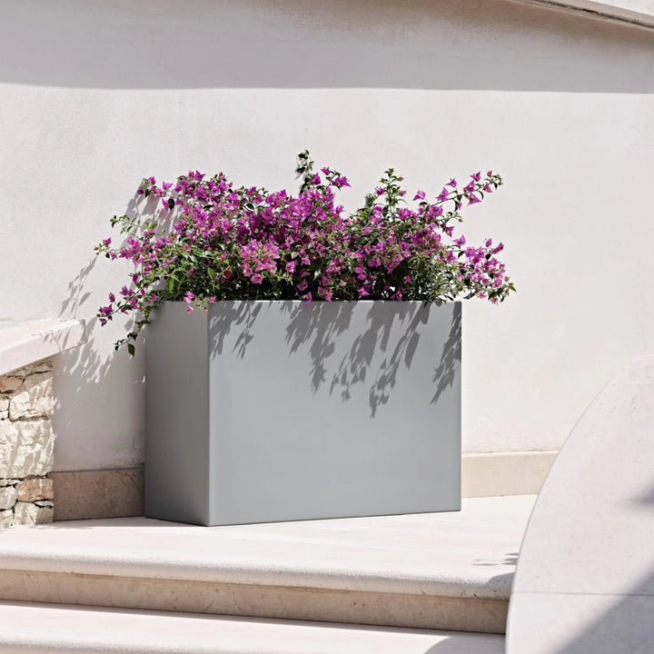 Fioriera rettangolare grigia con bouganvillea fucsia in fiore, posizionata su una scalinata esterna in pietra chiara.